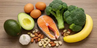 Alimentos saudáveis ​​que aumentam a testosterona sobre uma mesa rústica de madeira: abacate, ovos, salmão, nozes, banana, brócolis e um bulbo de alho.